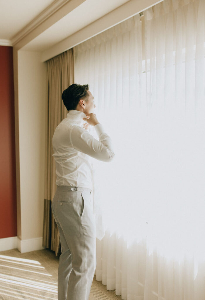 groom putting on his tie while looking out the window in the getting ready room of the hotel