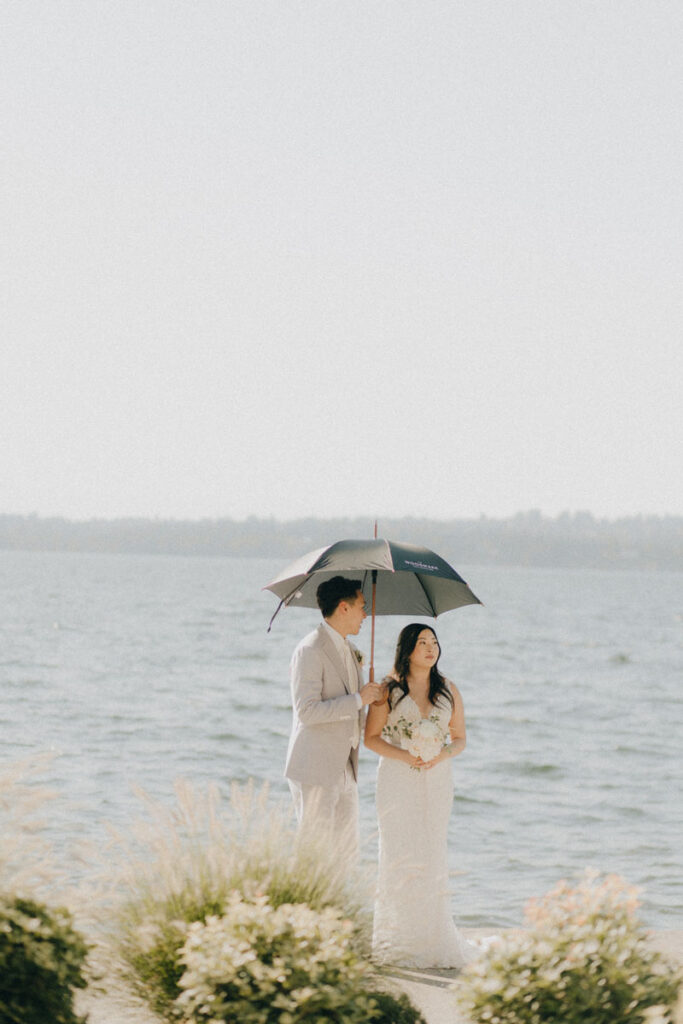 groom holding umbrella for bride on a hot sunny day in front of the water 